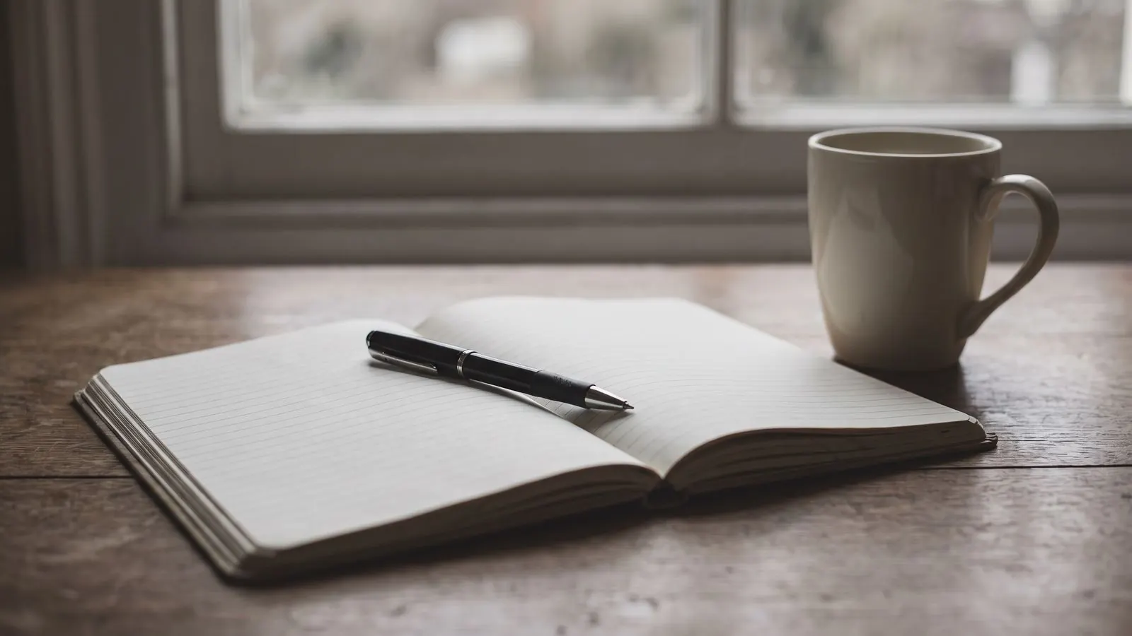 An open ruled notebook on a desk with a pen laid across the page and a mug beside it