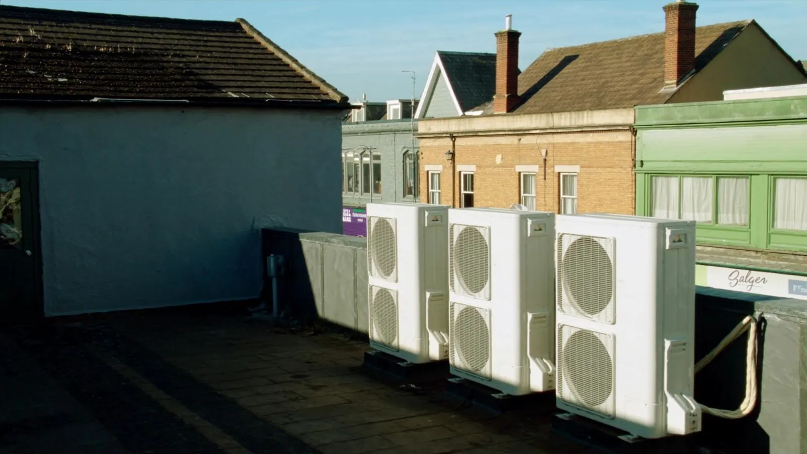 A row of commercial air conditioning condensers on a dry flat roof above British shopfronts on a clear day
